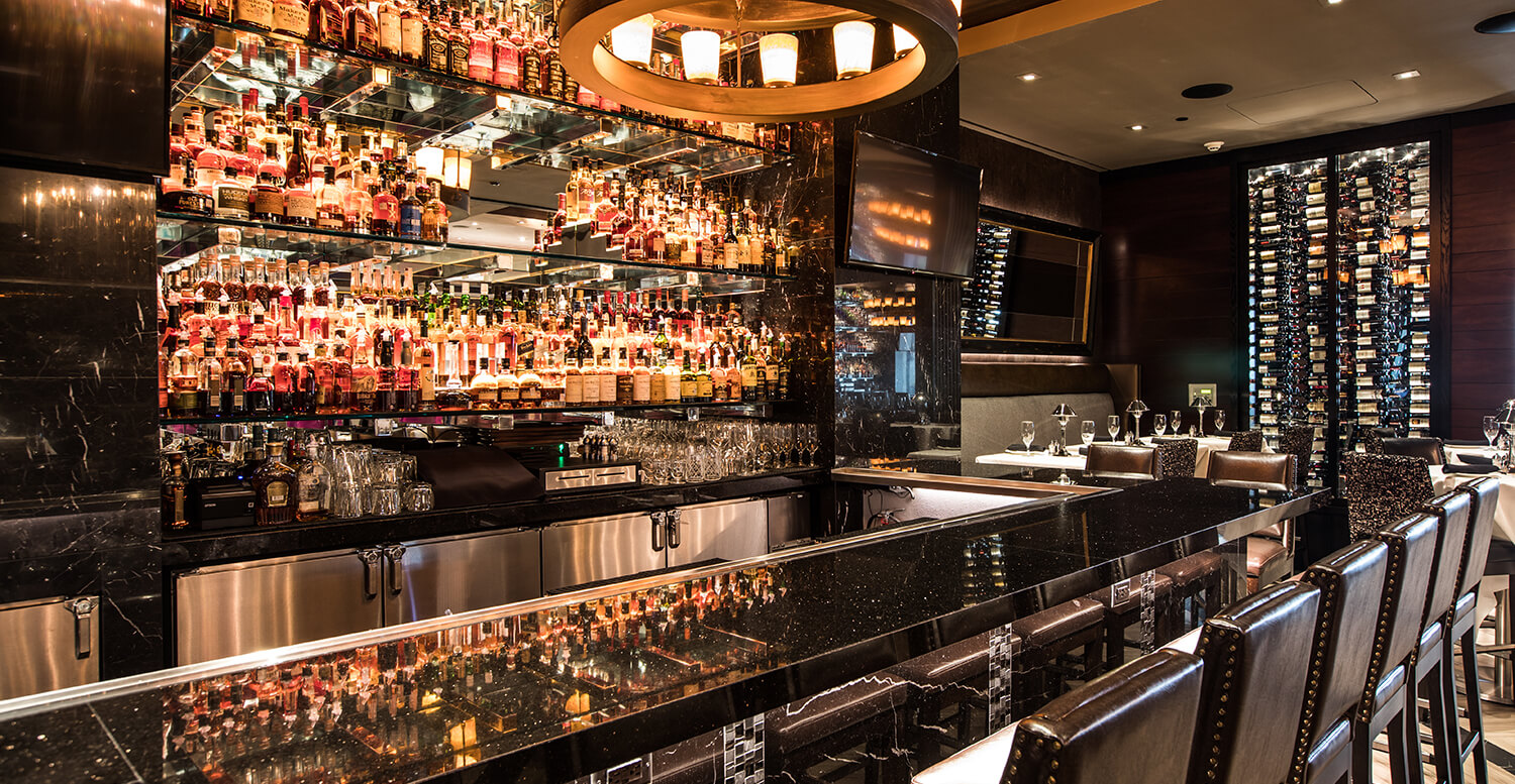 A luxurious bar with a black marble countertop, stools, and shelves stocked with various bottles of alcohol, along with a chandelier and wine storage.